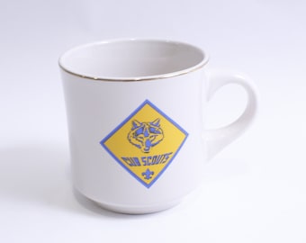 Puede incluir: Taza de cerámica blanca con un borde dorado. La taza presenta un logotipo azul y amarillo de los Boy Scouts con una cabeza de lobo y el texto "Cub Scouts".