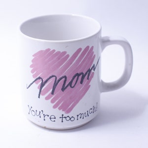 Pode incluir: Caneca de cerâmica branca com um desenho de coração rosa e a palavra "Mãe" escrita em preto dentro do coração. O texto "Você é demais!" está escrito abaixo do coração.