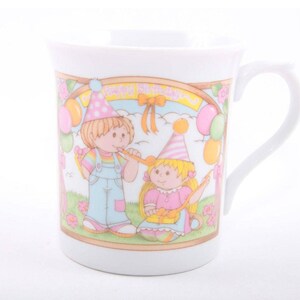 May include: A white ceramic mug featuring a colourful illustration of two children wearing party hats and holding noisemakers. The scene includes balloons, gifts, and the text Happy Birthday in a decorative frame against a grassy background.