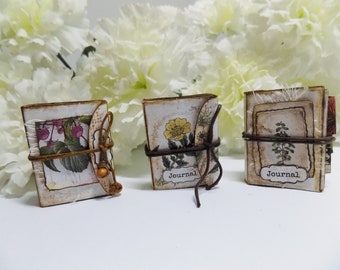 May include: Three small, handmade journals with floral designs and the word "Journal" on the front. The journals are tied with twine and have a rustic, vintage aesthetic.
