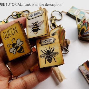 May include: Four miniature books with bee illustrations and titles "Queen", "Journal", "Notes", and "Nature" on a yellow background. The books are held in a hand.