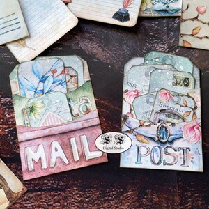 May include: Two decorative mail and post envelopes with a vintage aesthetic. The envelopes are decorated with floral patterns, including magnolia blossoms, and have a layered, dimensional look. The envelopes are labeled "MAIL" and "POST" in a distressed font.