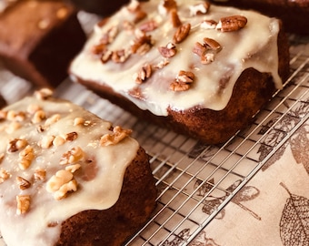 May include: A close-up of a cooling rack with several loaves of banana bread topped with a white glaze and pecans. The bread is on a beige tablecloth with a floral pattern.