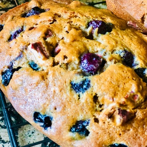 May include: A close-up of a loaf of blueberry bread with pecans. The bread is golden brown and has a slightly crispy crust. The blueberries are scattered throughout the bread and some are visible on the surface.