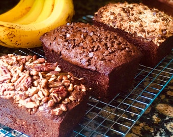 May include: Three loaves of banana bread on a wire rack. The bread on the left is topped with pecans, the middle loaf is topped with chocolate chips, and the loaf on the right is topped with shredded coconut.
