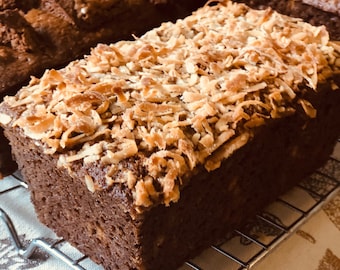May include: A close-up of a loaf of chocolate coconut bread on a wire rack. The bread is topped with shredded coconut.