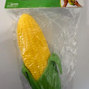 May include: A yellow corn-shaped squeeze toy with green husks at the base, packaged in a clear plastic bag with a header card labeled Squeezy. The packaging includes a warning about small parts and a graphic showing the toy being compressed.