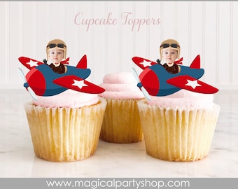 May include: Two red, white, and blue airplane cupcake toppers with a baby pilot wearing a brown aviator hat and goggles. The text "Cupcake Toppers" is at the top of the image.