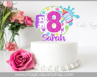 May include: A pink and purple glitter cake topper with the number 8, the name Sarah, and images of slime and glitter glue. The topper is on a white cake with white frosting. The text "Cake Topper" is visible in the background.