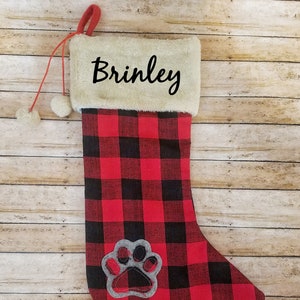 May include: Red and black buffalo plaid Christmas stocking with a white faux fur cuff. The stocking has a gray paw print applique and is personalized with the name "Brinley".