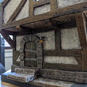 May include: A miniature model of a small, white building with a wooden door and a stone archway. The building has a brown wooden roof and a stone foundation. There is a small light on the wall next to the door.