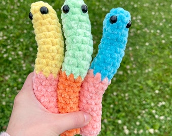 May include: Three crocheted worm plushies in different colors: yellow, green, and blue. Each worm has black eyes and a contrasting colored tail.