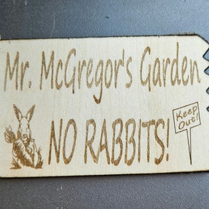 以下が含まれることがあります： 「Mr. McGregor's Garden No Rabbits! 」と書かれた木製の看板と、ニンジンを持った漫画のウサギ。看板には「Keep Out!」と書かれた小さな看板も付いています。