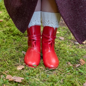 May include: A pair of red leather boots with laces, worn with white lace socks. The boots are on a bed of green moss.