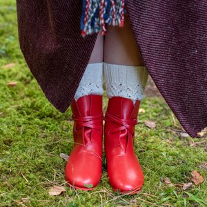 May include: A pair of red leather boots with laces tied at the ankle. The boots are worn by a person standing on a bed of green moss.