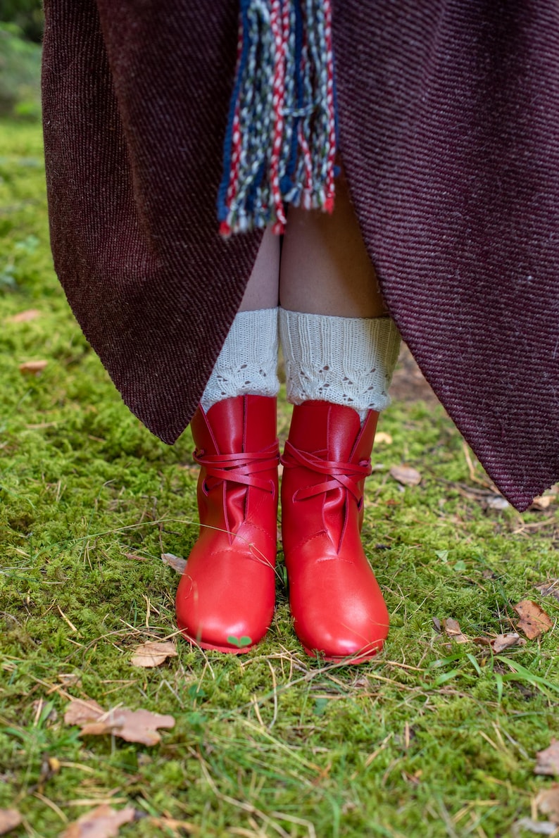 May include: A pair of red leather boots with laces tied at the ankle. The boots are worn by a person standing on a bed of green moss.