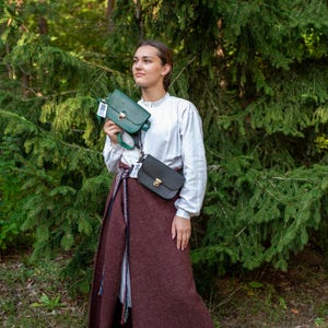 May include: A woman wearing a white shirt and a long brown skirt is standing in a forest. She is holding two leather purses, one green and one black.