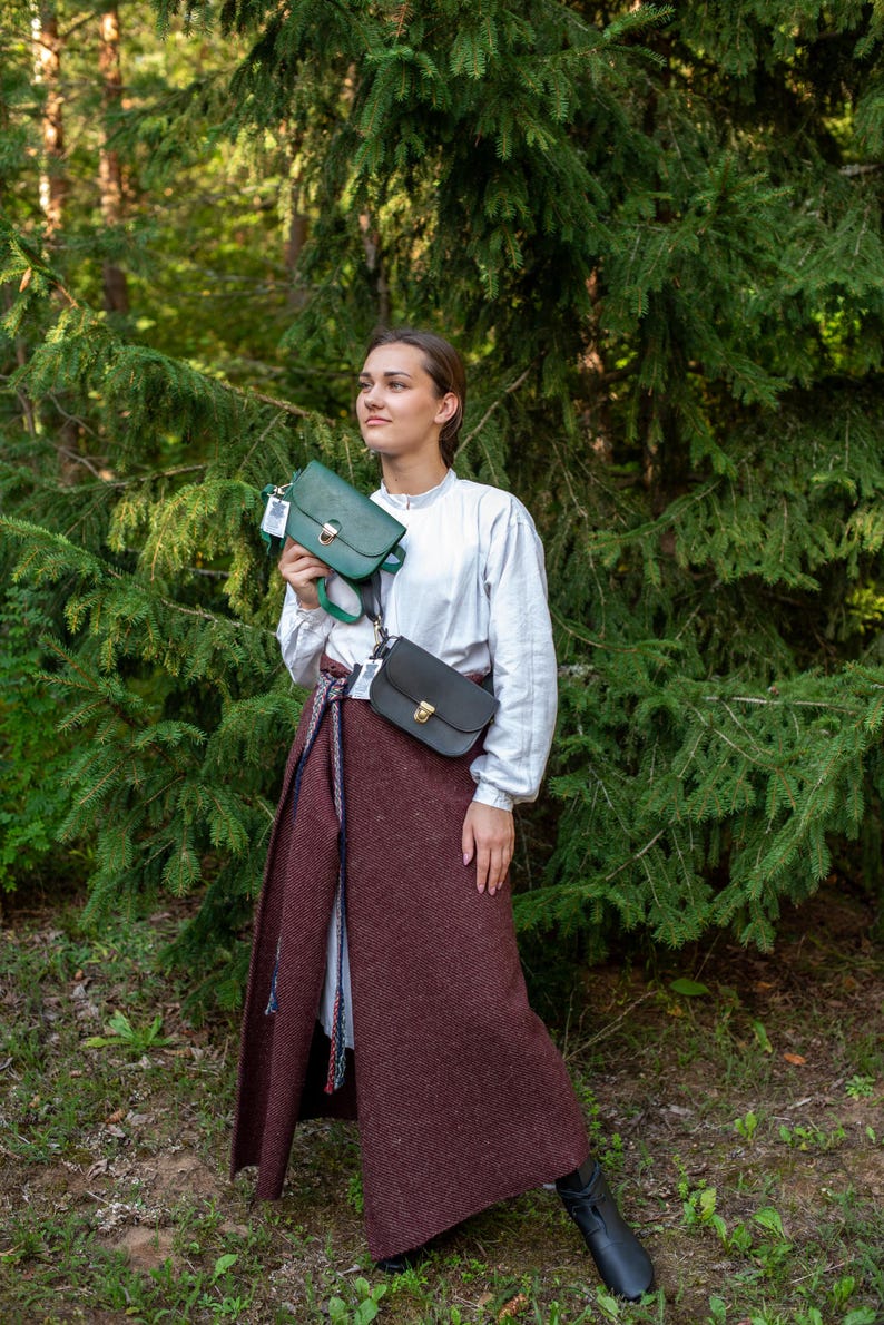 May include: A woman wearing a white shirt and a long brown skirt is standing in a forest. She is holding two leather purses, one green and one black.
