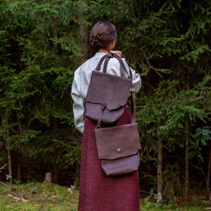 Może przedstawiać: Osoba w jasnej koszuli i bordowej sp&oacute;dnicy niesie dwie brązowe sk&oacute;rzane torby z paskami na ramię. Torby mają rustykalną, vintage estetykę, odpowiednią do przenoszenia przedmiot&oacute;w. Tło przedstawia bujny zielony las.