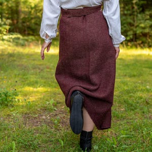 May include: A person wearing a white blouse and a long, burgundy skirt walks through a grassy area. The skirt has a subtle herringbone pattern and the person is wearing black boots.