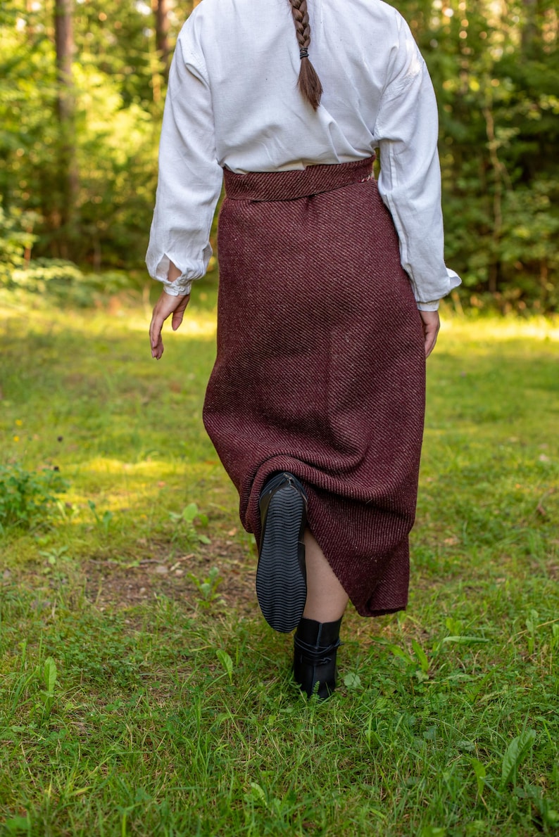 May include: A person wearing a white blouse and a long, burgundy skirt walks through a grassy area. The skirt has a subtle herringbone pattern and the person is wearing black boots.