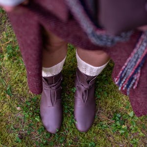 May include: A pair of brown leather lace-up boots with white socks. The boots are worn by a person standing on a bed of green moss.