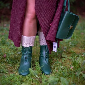 May include: A pair of dark green leather boots with laces tied around the ankles. The boots are worn by a person wearing a burgundy skirt and white socks. The person is standing on a bed of green moss.