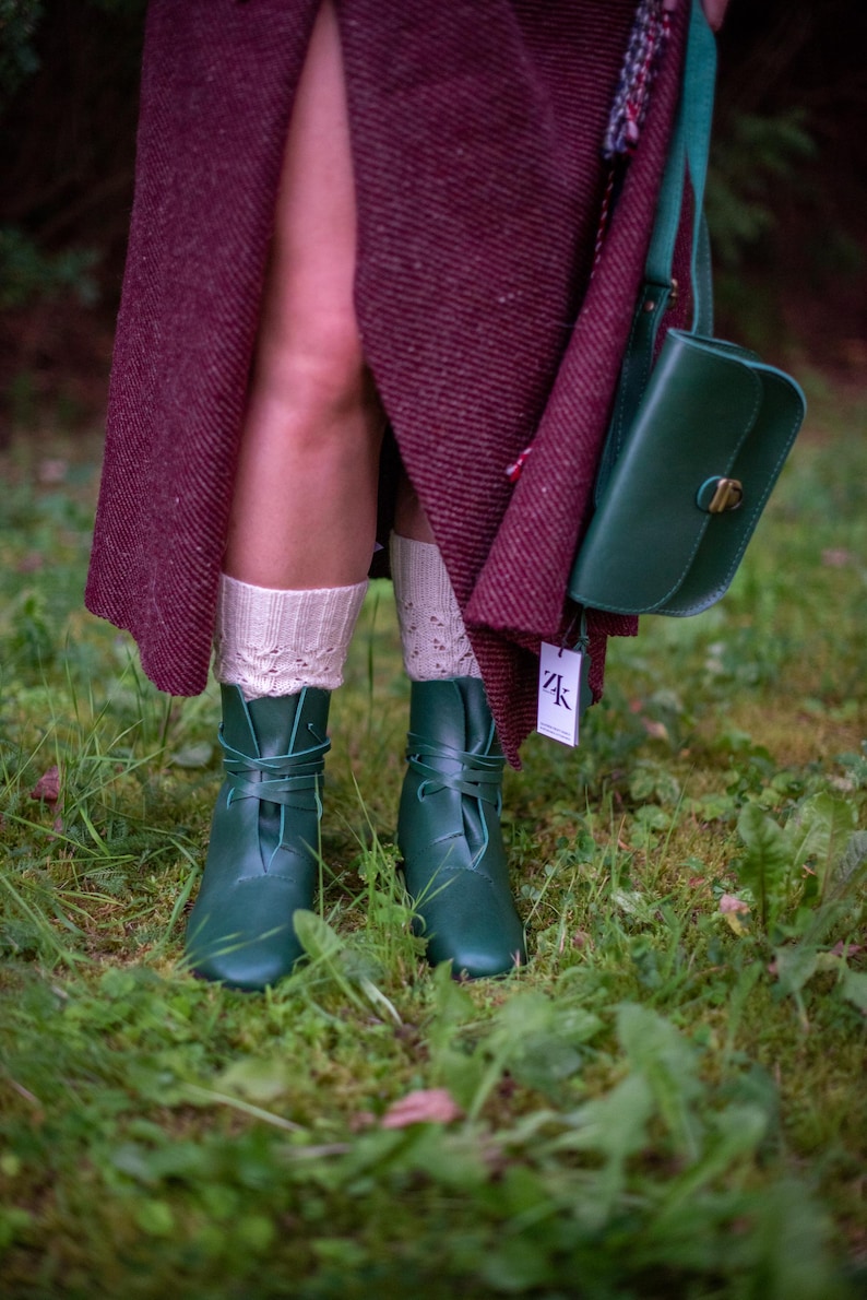 May include: A pair of dark green leather boots with laces tied around the ankles. The boots are worn by a person wearing a burgundy skirt and white socks. The person is standing on a bed of green moss.