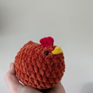 Puede incluir: Un juguete de pollo de peluche hecho a mano en tonos naranja, rojo y amarillo. El cuerpo es naranja texturizado, con una cresta roja y un pico amarillo. El pollo tiene un ojo negro y est&aacute; dise&ntilde;ado para ser suave y tierno.