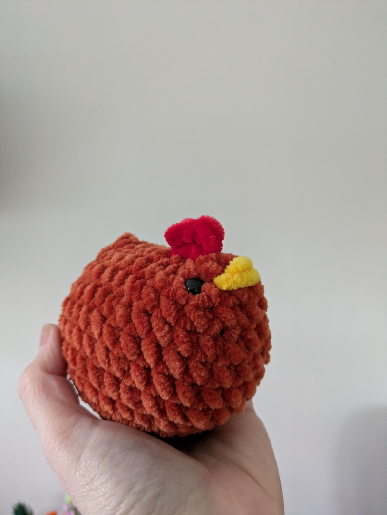 Puede incluir: Un juguete de pollo de peluche hecho a mano en tonos naranja, rojo y amarillo. El cuerpo es naranja texturizado, con una cresta roja y un pico amarillo. El pollo tiene un ojo negro y est&aacute; dise&ntilde;ado para ser suave y tierno.