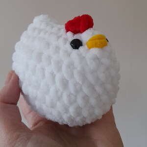 Puede incluir: Un juguete de pollo de peluche blanco hecho a mano, con una cresta roja, ojos negros y un pico amarillo. El pollo suave y redondo se sostiene en una mano, mostrando su textura esponjosa y su dise&ntilde;o caprichoso. Este adorable juguete es perfecto para ni&ntilde;os.