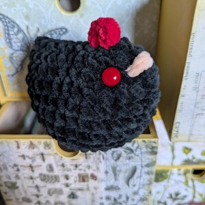 Puede incluir: Una criatura negra de ganchillo hecha a mano con un pomp&oacute;n rojo en la parte superior, un ojo de bot&oacute;n rojo y una peque&ntilde;a oreja beige. El peluche est&aacute; en un caj&oacute;n decorativo con ilustraciones bot&aacute;nicas y un dise&ntilde;o de mariposa.