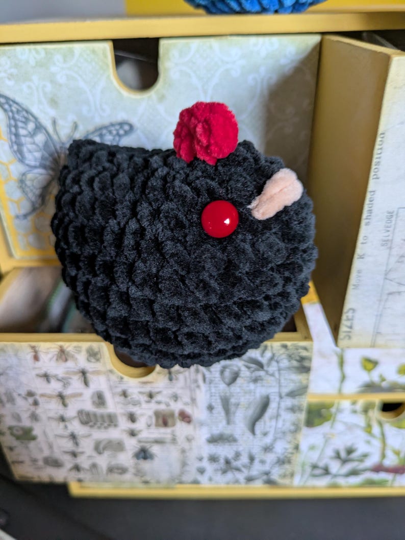Puede incluir: Una criatura negra de ganchillo hecha a mano con un pomp&oacute;n rojo en la parte superior, un ojo de bot&oacute;n rojo y una peque&ntilde;a oreja beige. El peluche est&aacute; en un caj&oacute;n decorativo con ilustraciones bot&aacute;nicas y un dise&ntilde;o de mariposa.