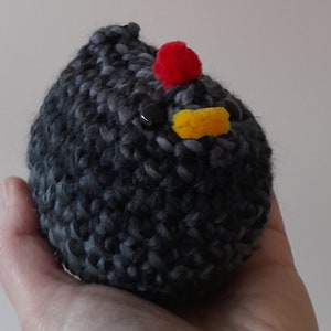 Puede incluir: Un pollo hecho a mano, tejido a ganchillo, con un cuerpo gris oscuro y negro. El pollo tiene una cresta roja, un pico amarillo y ojos negros de bot&oacute;n. El peluche se sostiene en una mano.
