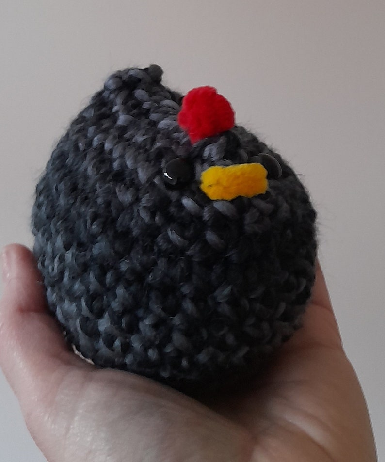 Puede incluir: Un pollo hecho a mano, tejido a ganchillo, con un cuerpo gris oscuro y negro. El pollo tiene una cresta roja, un pico amarillo y ojos negros de bot&oacute;n. El peluche se sostiene en una mano.