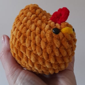 Puede incluir: Un juguete de pollo de peluche hecho a mano en un c&aacute;lido tono naranja. El pollo presenta una cresta roja, ojos negros y un pico amarillo. El material suave y texturizado le da al juguete una apariencia tierna. Este art&iacute;culo artesanal es probablemente un juguete para ni&ntilde;os o un objeto decorativo.