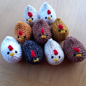 Puede incluir: Una colecci&oacute;n de diez adornos de pollo de ganchillo en varios colores, incluyendo blanco, marr&oacute;n y gris. Cada pollo tiene una cresta roja, ojos negros y un pico amarillo. Los adornos est&aacute;n dispuestos sobre una superficie de madera.