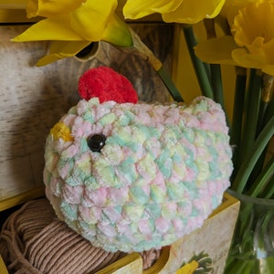 Puede incluir: Un pollo de ganchillo hecho a mano en colores pastel rosa, verde y amarillo, con una cresta roja y un ojo negro. El pollo est&aacute; colocado en una caja de madera con una madeja de hilo marr&oacute;n, con narcisos amarillos en el fondo.