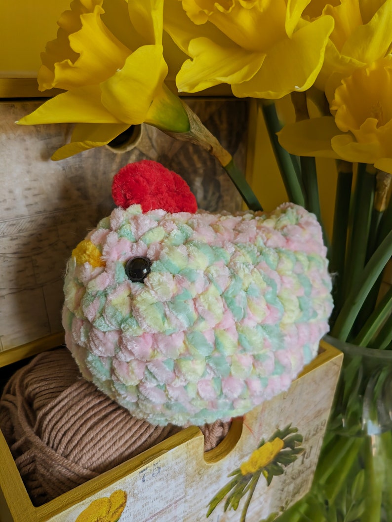 Puede incluir: Un pollo de ganchillo hecho a mano en colores pastel rosa, verde y amarillo, con una cresta roja y un ojo negro. El pollo est&aacute; colocado en una caja de madera con una madeja de hilo marr&oacute;n, con narcisos amarillos en el fondo.