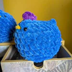 Puede incluir: Un pollo de crochet azul con una cresta morada y un pico amarillo. El animal de peluche hecho a mano tiene un cuerpo redondo y est&aacute; colocado sobre una caja de madera decorativa. El fondo es amarillo liso.