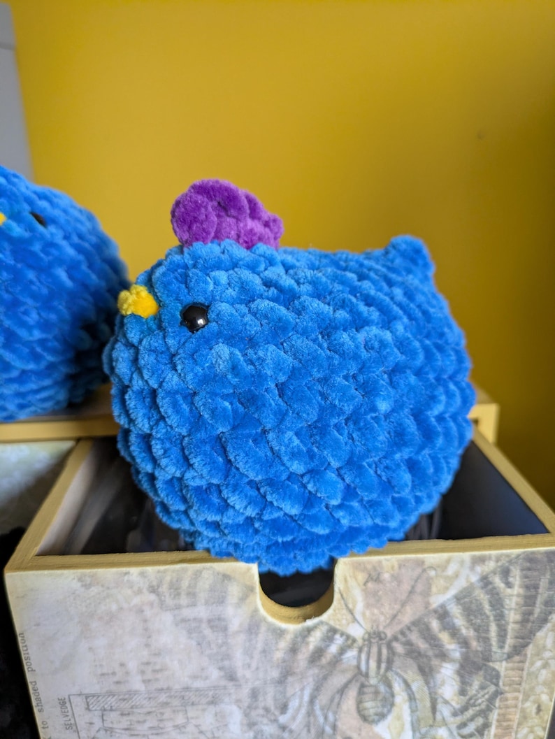 Puede incluir: Un pollo de crochet azul con una cresta morada y un pico amarillo. El animal de peluche hecho a mano tiene un cuerpo redondo y est&aacute; colocado sobre una caja de madera decorativa. El fondo es amarillo liso.
