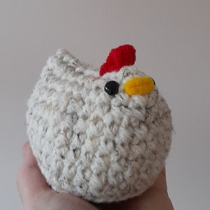 Puede incluir: Un juguete de pollo hecho a mano, tejido a ganchillo. El pollo es blanco con motas negras y grises, una cresta roja, un pico amarillo y un ojo negro. El juguete se sostiene en una mano sobre un fondo liso.