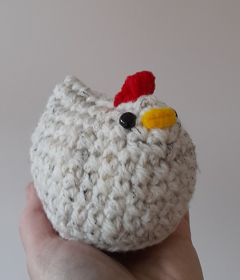 Puede incluir: Un juguete de pollo hecho a mano, tejido a ganchillo. El pollo es blanco con motas negras y grises, una cresta roja, un pico amarillo y un ojo negro. El juguete se sostiene en una mano sobre un fondo liso.