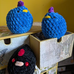 Puede incluir: Tres peluches hechos a mano con ganchillo. Dos son azules con detalles amarillos y morados, parecidos a p&aacute;jaros. El tercero es negro con detalles rojos y beige, posiblemente una criatura. Est&aacute;n sobre una caja de almacenamiento decorativa con estampado bot&aacute;nico.