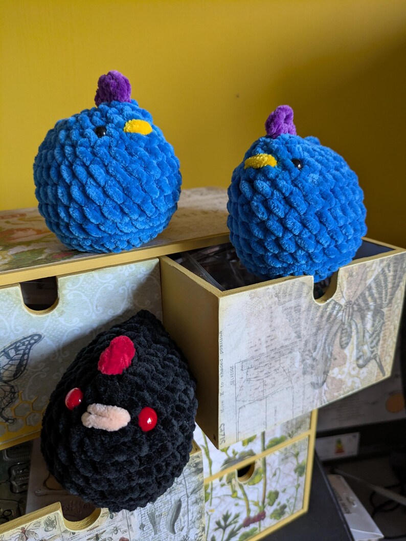 Puede incluir: Tres peluches hechos a mano con ganchillo. Dos son azules con detalles amarillos y morados, parecidos a p&aacute;jaros. El tercero es negro con detalles rojos y beige, posiblemente una criatura. Est&aacute;n sobre una caja de almacenamiento decorativa con estampado bot&aacute;nico.