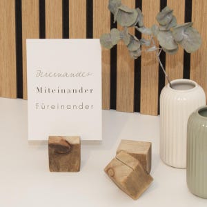 Könnte beinhalten: Eine weiße Karte mit den Worten "Beieinander, Miteinander, Füreinander" in schwarzer Schrift wird von einem kleinen Holzblock gehalten. Zwei weitere Holzblöcke und zwei Keramikvasen sind ebenfalls vorhanden. Der Hintergrund ist eine Holzwand.