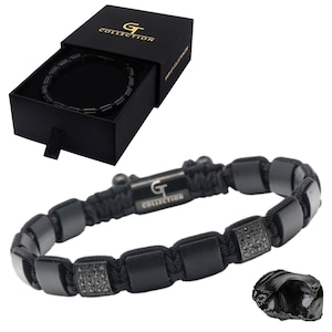 Op de afbeelding: Een zwarte armband met vierkante kralen en een gevlochten koord, gepresenteerd in een zwarte doos. De armband heeft een metalen sluiting met het "GT COLLECTION" logo. Een zwarte steen is ook zichtbaar.