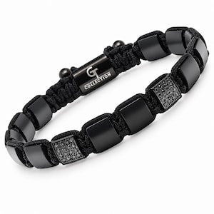 Op de afbeelding: Een zwarte armband van gevlochten koord met gladde vierkante kralen en twee vierkante kralen ingelegd met kleine zwarte steentjes. Een centrale rechthoekige sluiting van zwart metaal toont de witte tekst GT COLLECTION, geflankeerd door twee zwarte kralen.