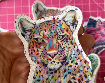 May include: A colourful sticker depicting a leopard's face, with vibrant blues, greens, yellows, and pinks. The sticker has a white border and is held by a hand. Other stickers are visible in the background.