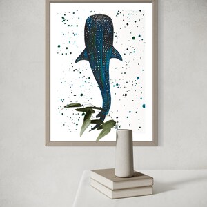 May include: Framed art print featuring a blue whale shark illustration with white spots and a dark blue body. The artwork has a white background with teal paint splatters and green leaves. The frame is a light brown color.